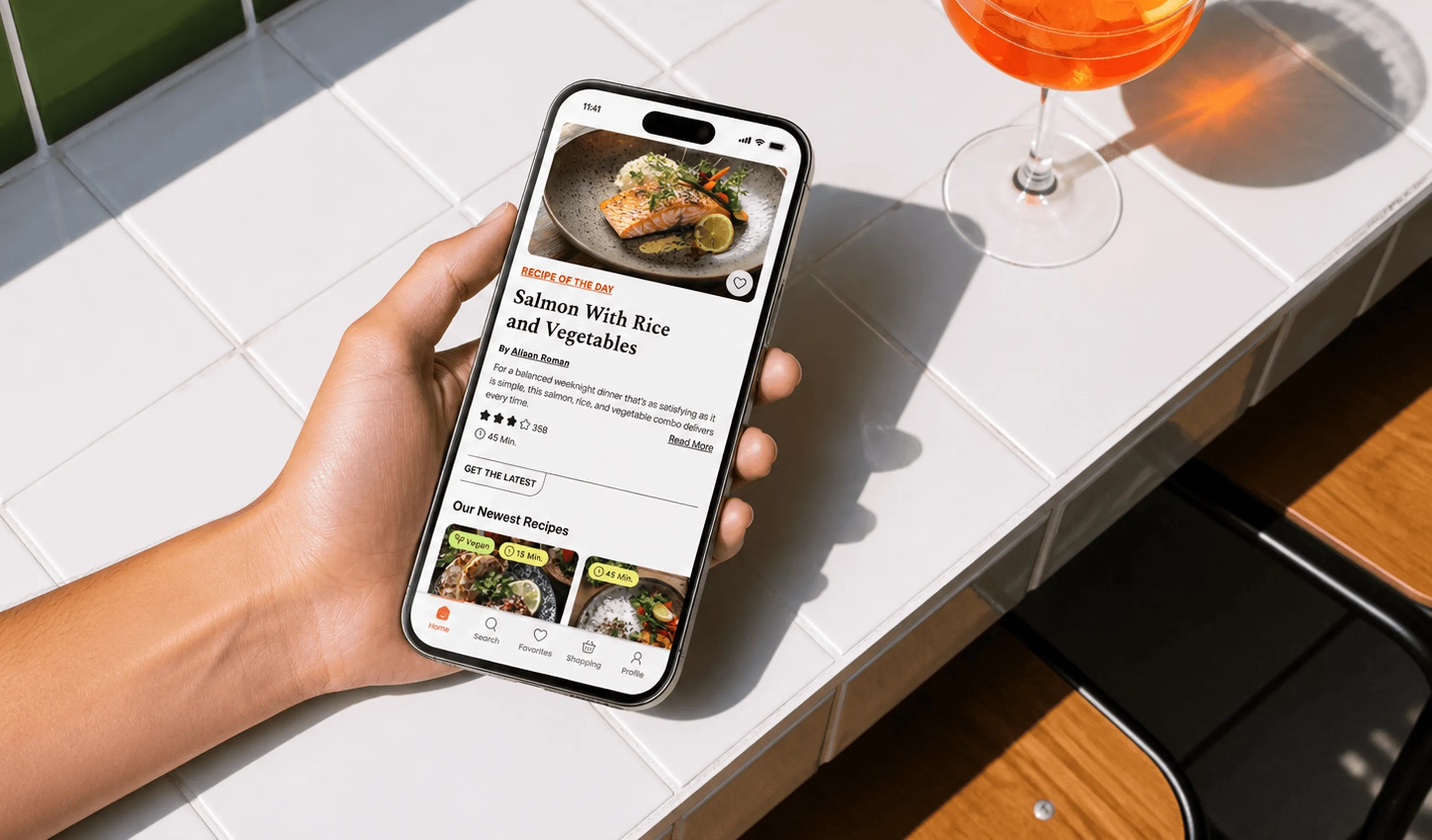 Dishcovery Home screen with Recipe of the Day on a phone held over a white tiled table